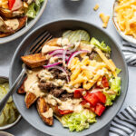 A bowl with lettuce, tomato, pickles, red onion, cheddar cheese, ground beef, potato wedges, and a creamy sauce, surrounded by similar ingredients and sauce bowls.