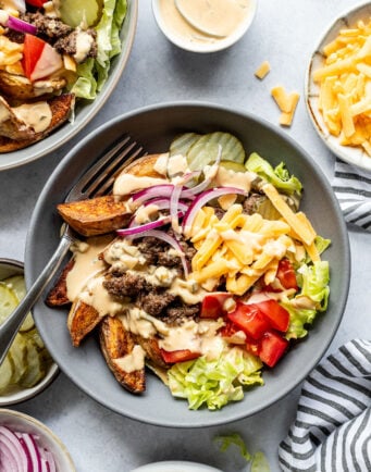 A bowl with lettuce, tomato, pickles, red onion, cheddar cheese, ground beef, potato wedges, and a creamy sauce, surrounded by similar ingredients and sauce bowls.