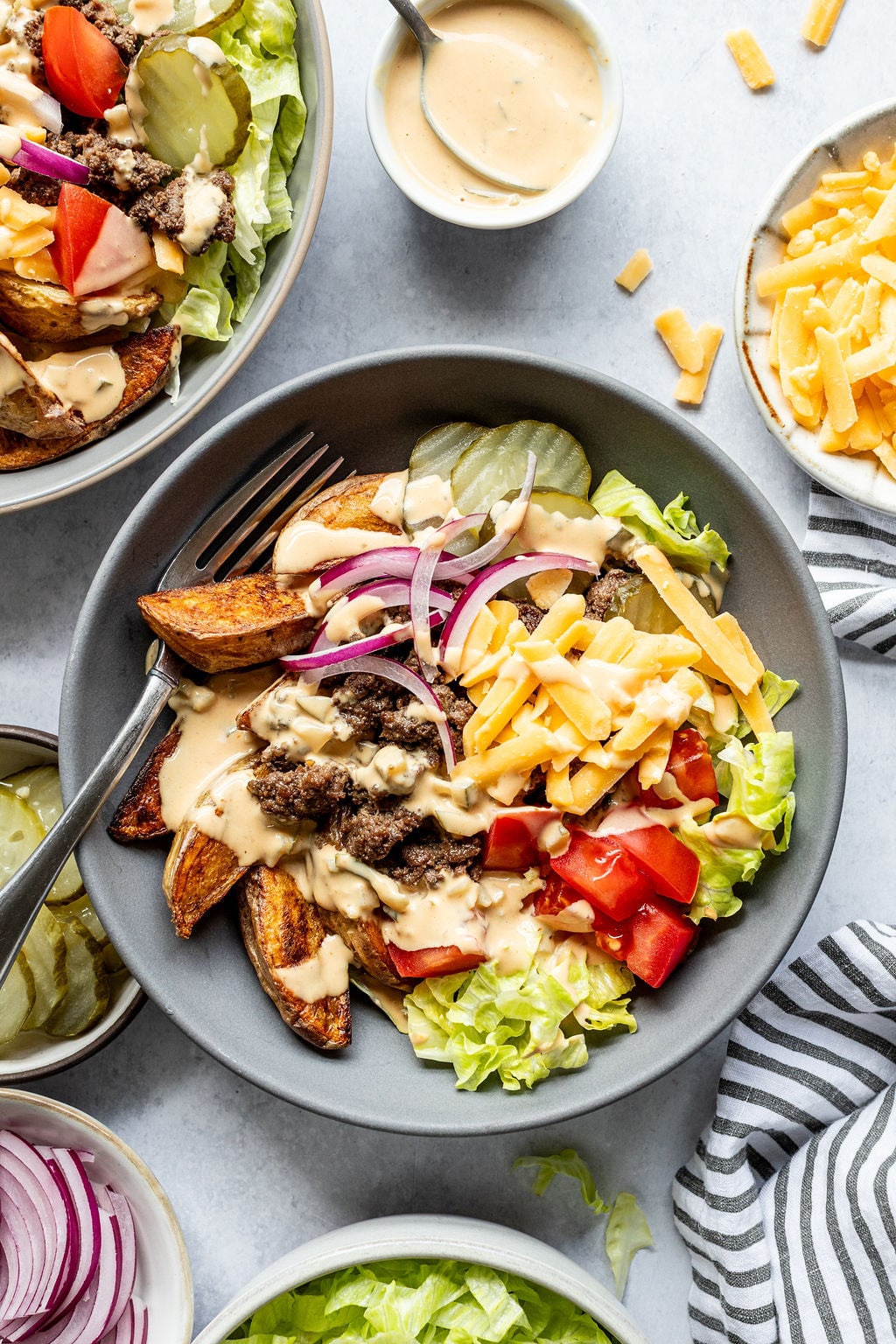 A bowl with lettuce, tomato, pickles, red onion, cheddar cheese, ground beef, potato wedges, and a creamy sauce, surrounded by similar ingredients and sauce bowls.