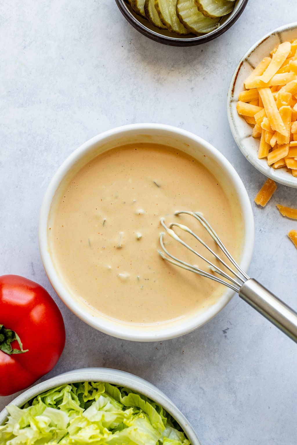 A bowl of creamy sauce with a whisk, surrounded by bowls of sliced pickles, shredded cheddar cheese, chopped lettuce, and a tomato.
