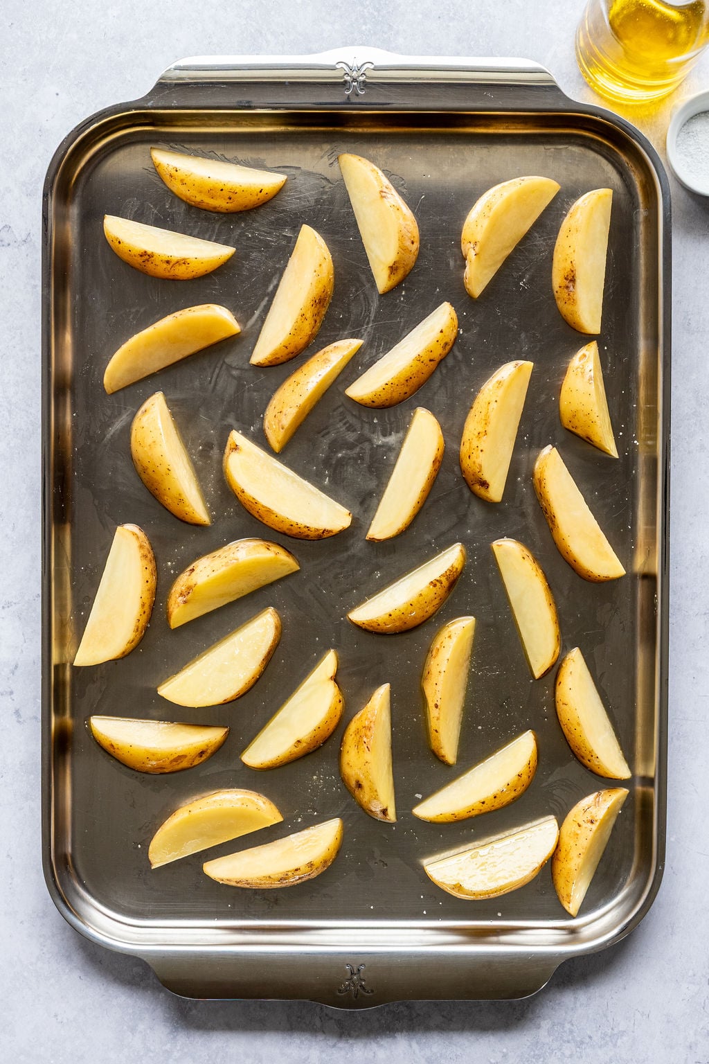 Raw potato wedges are evenly spaced on a metal baking sheet, ready to be baked. A bottle of oil and a small bowl of salt are visible in the upper right corner.