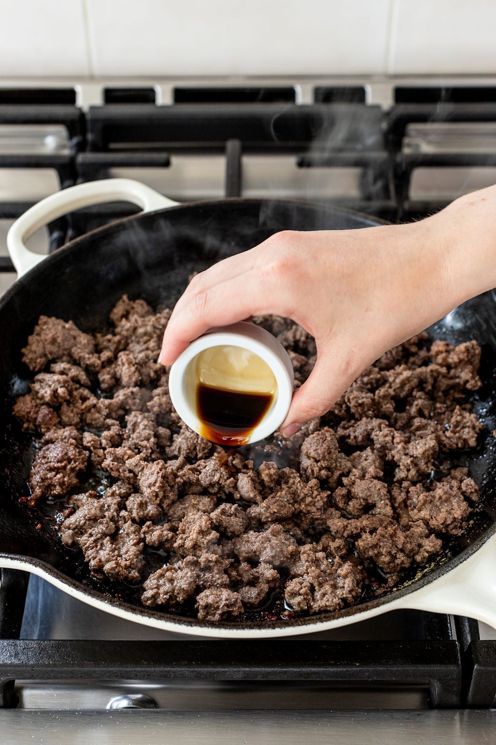 A hand pours a small cup of dark liquid into a skillet of cooked ground beef on a stovetop.