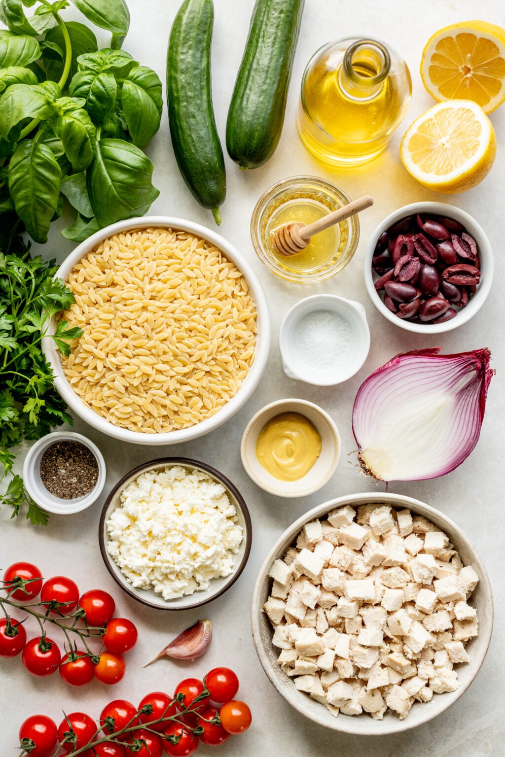 Top-down view of orzo pasta, feta cheese, cubed chicken, tomatoes, cucumbers, olives, basil, parsley, red onion, olive oil, lemon, mustard, honey, garlic, salt, and pepper on a white surface.