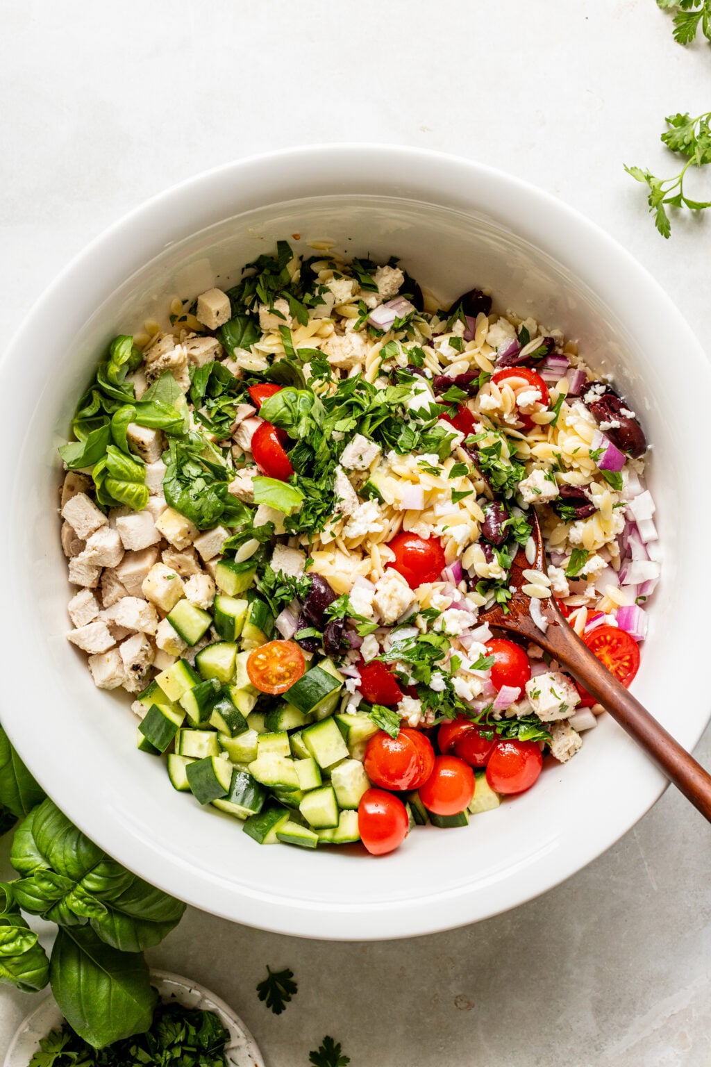 Orzo pasta salad with chicken, cucumber, tomatoes, feta, olives, and herbs in a white bowl.