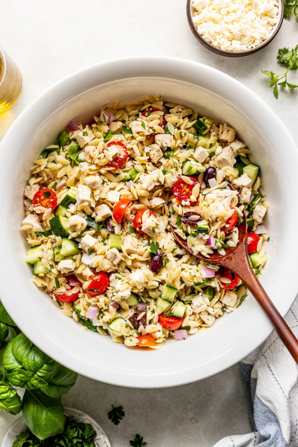 A white bowl filled with orzo pasta salad containing diced chicken, cherry tomatoes, cucumbers, red onion, olives, and feta cheese, with a wooden spoon resting inside.
