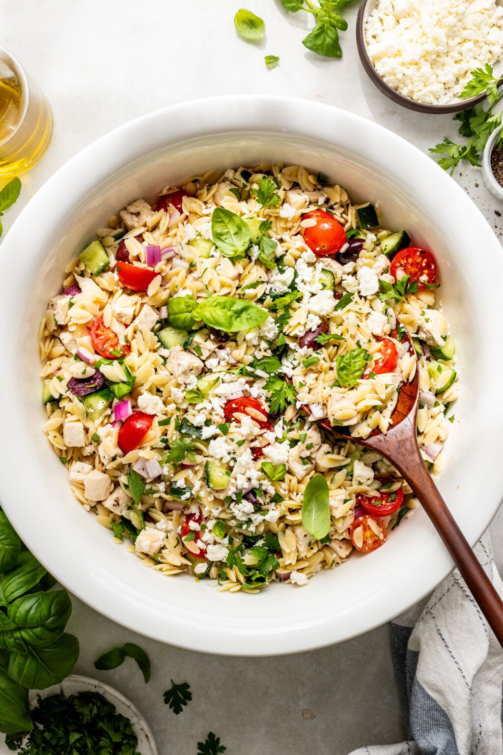 A white bowl filled with orzo pasta salad containing cherry tomatoes, cucumber, red onion, chicken, fresh herbs, and crumbled feta cheese, with a wooden serving spoon.