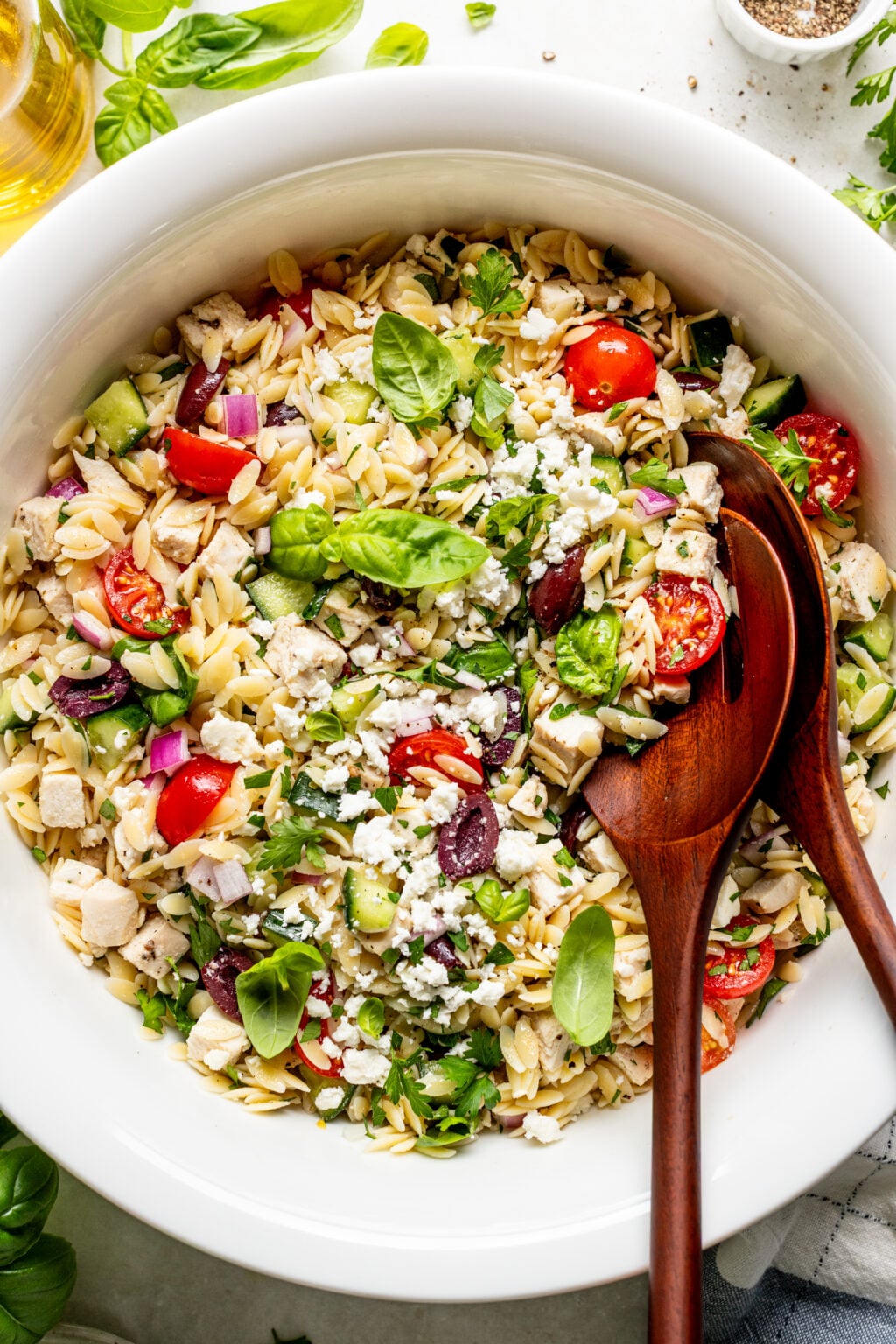 A white bowl filled with orzo pasta salad, featuring cherry tomatoes, feta cheese, olives, diced chicken, red onions, cucumber, herbs, and wooden serving utensils.