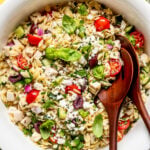 A white bowl filled with orzo pasta salad, featuring cherry tomatoes, feta cheese, olives, diced chicken, red onions, cucumber, herbs, and wooden serving utensils.