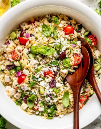 A white bowl filled with orzo pasta salad, featuring cherry tomatoes, feta cheese, olives, diced chicken, red onions, cucumber, herbs, and wooden serving utensils.