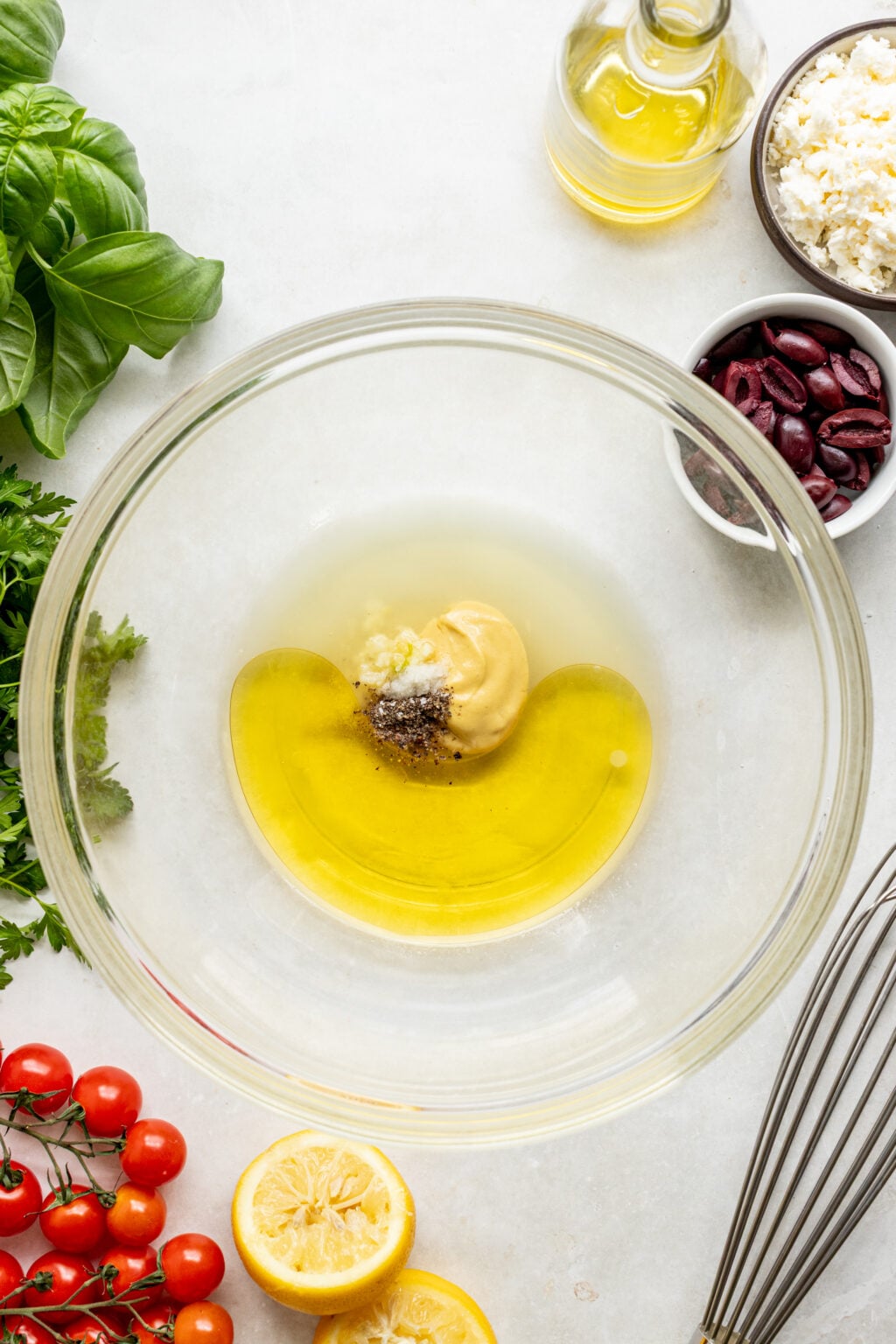 A glass bowl contains olive oil, mustard, garlic, salt, and pepper, surrounded by fresh basil, parsley, cherry tomatoes, lemon, olives, feta cheese, and a whisk.
