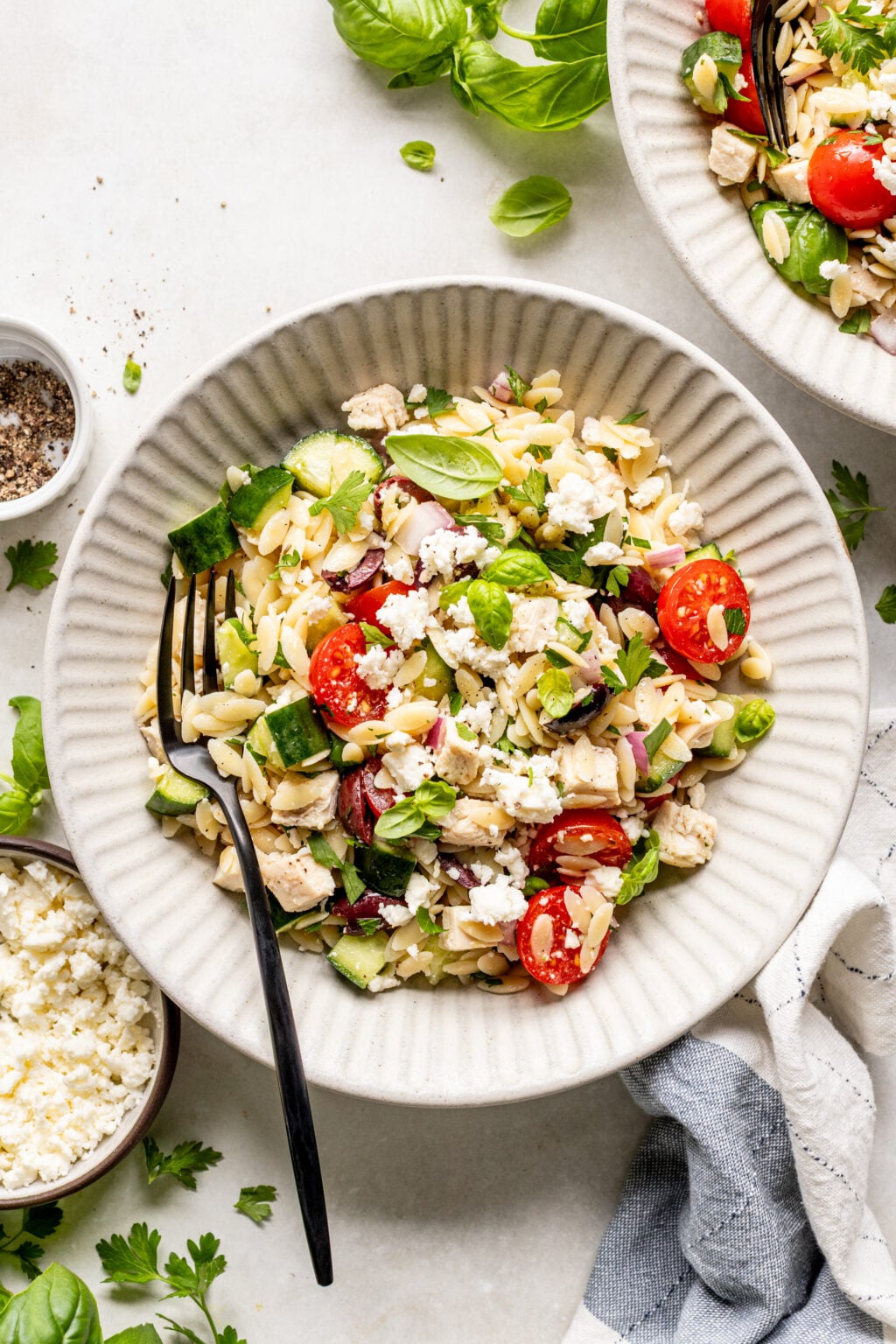 A bowl of orzo pasta salad with cherry tomatoes, cucumber, red onion, spinach, grilled chicken, and crumbled feta cheese, served with a fork.