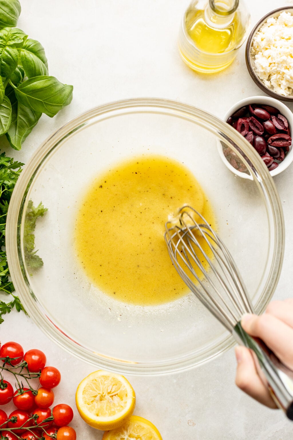 A hand whisks a yellow vinaigrette in a glass bowl, surrounded by fresh basil, parsley, cherry tomatoes, lemon halves, olives, feta cheese, and a bottle of oil.