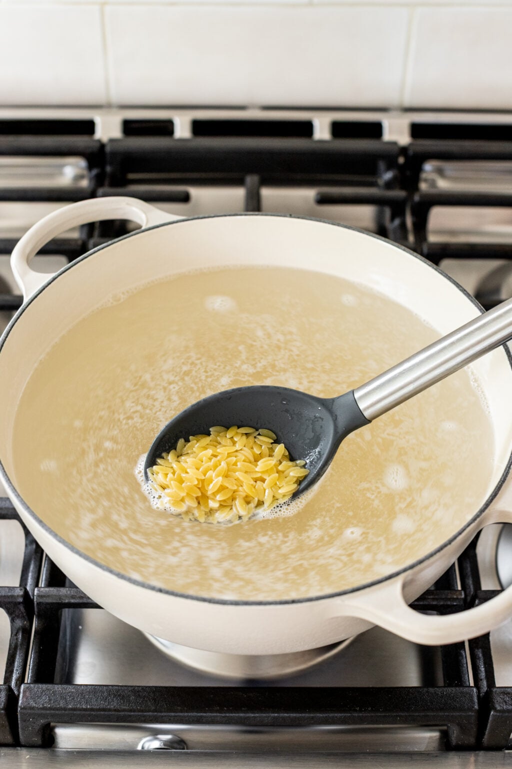 A pot of boiling water on a stovetop with a slotted spoon holding uncooked orzo pasta above the surface.