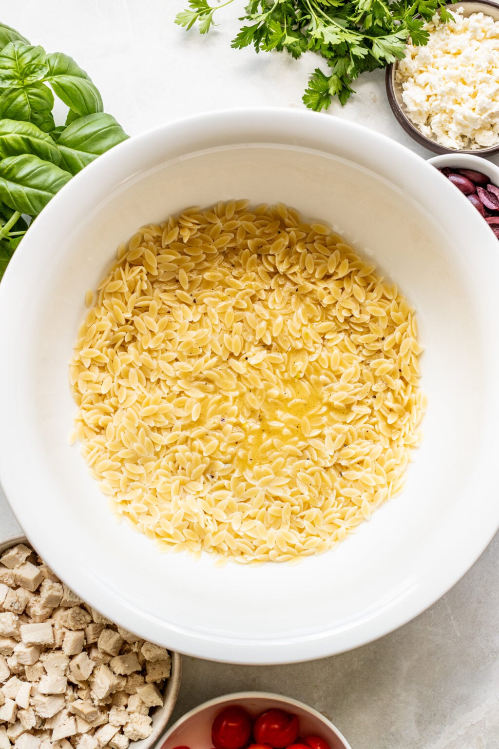 A white bowl filled with uncooked orzo pasta, surrounded by bowls of diced chicken, cherry tomatoes, feta cheese, olives, and fresh herbs.