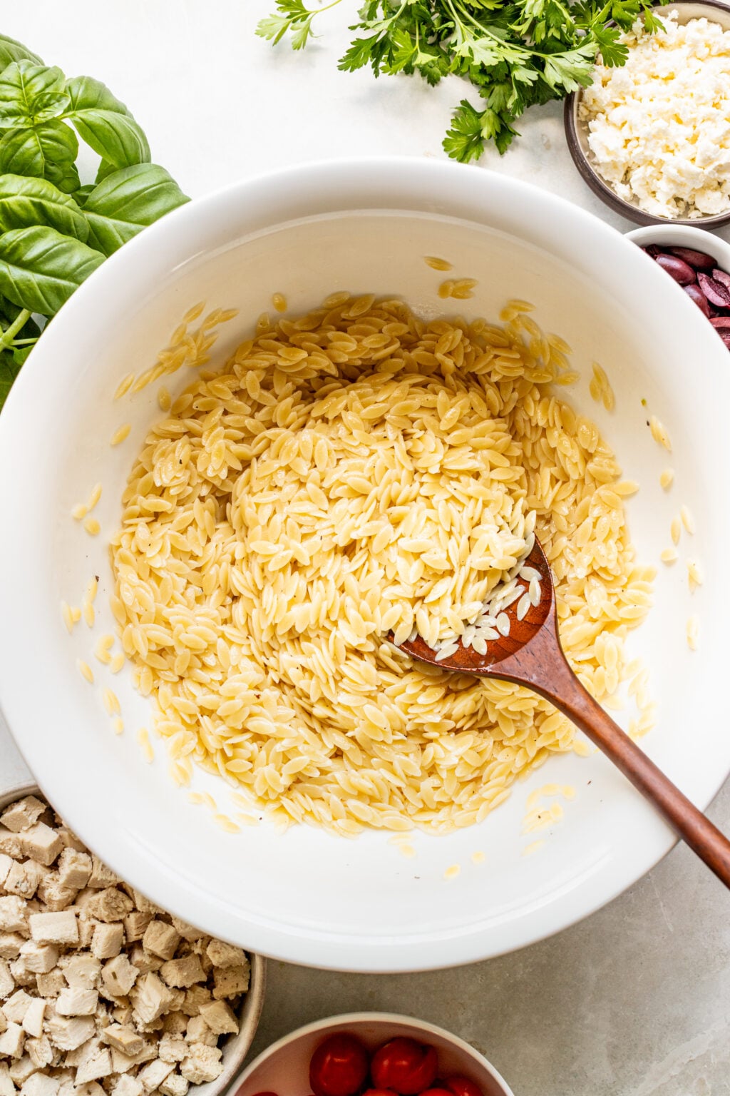 A large bowl of cooked orzo pasta with a wooden spoon, next to bowls of chopped chicken, feta cheese, olives, basil, and parsley.