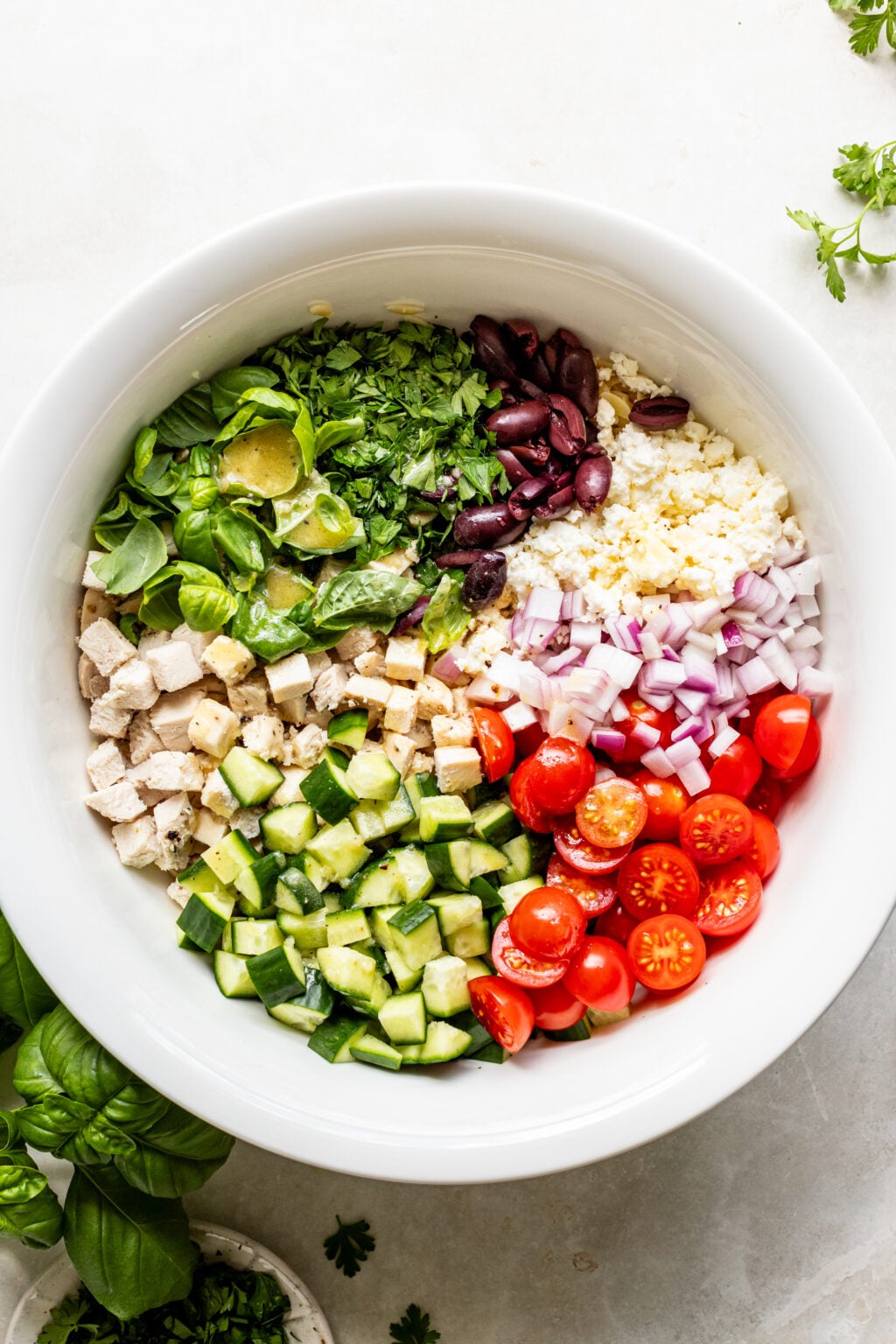A white bowl contains separate sections of chopped lettuce, olives, feta cheese, red onion, cherry tomatoes, cucumber, and cooked chicken.