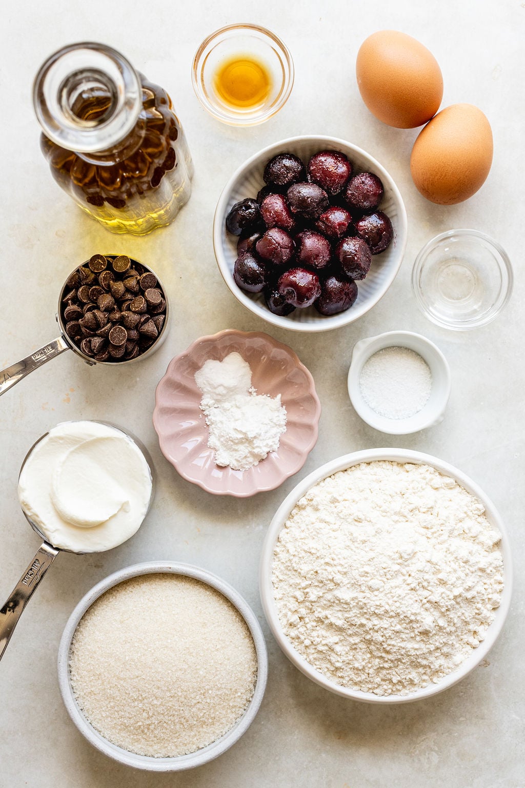 Baking ingredients arranged on a counter, including flour, sugar, eggs, chocolate chips, cherries, oil, vanilla, baking powder, salt, and yogurt in separate bowls and cups.