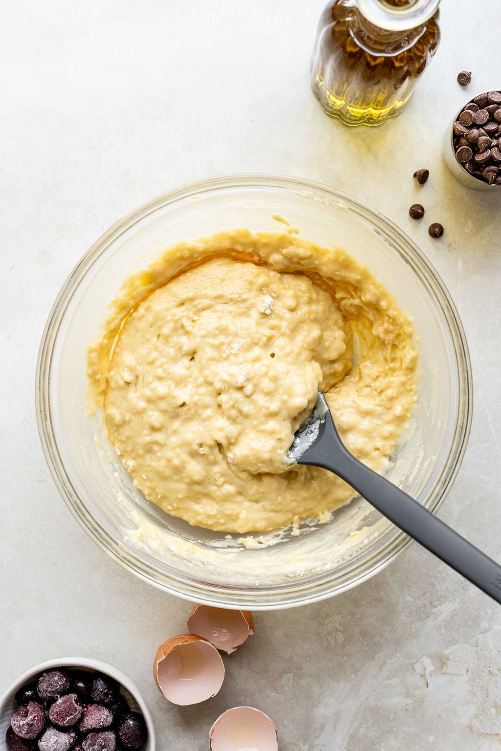 A glass bowl of muffin batter with a spatula, surrounded by chocolate chips, eggshells, and a bottle of oil on a light surface.