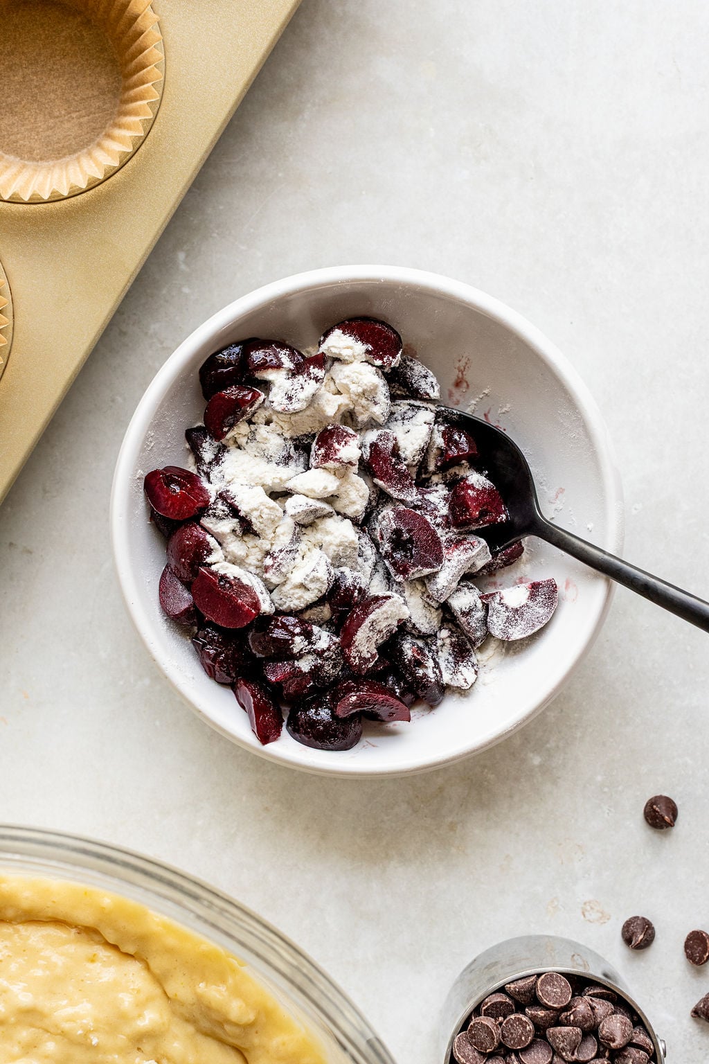 A bowl of halved cherries mixed with flour, with a black spoon inside. Nearby are a muffin tin with liners, a batter-filled bowl, and a small glass of chocolate chips.