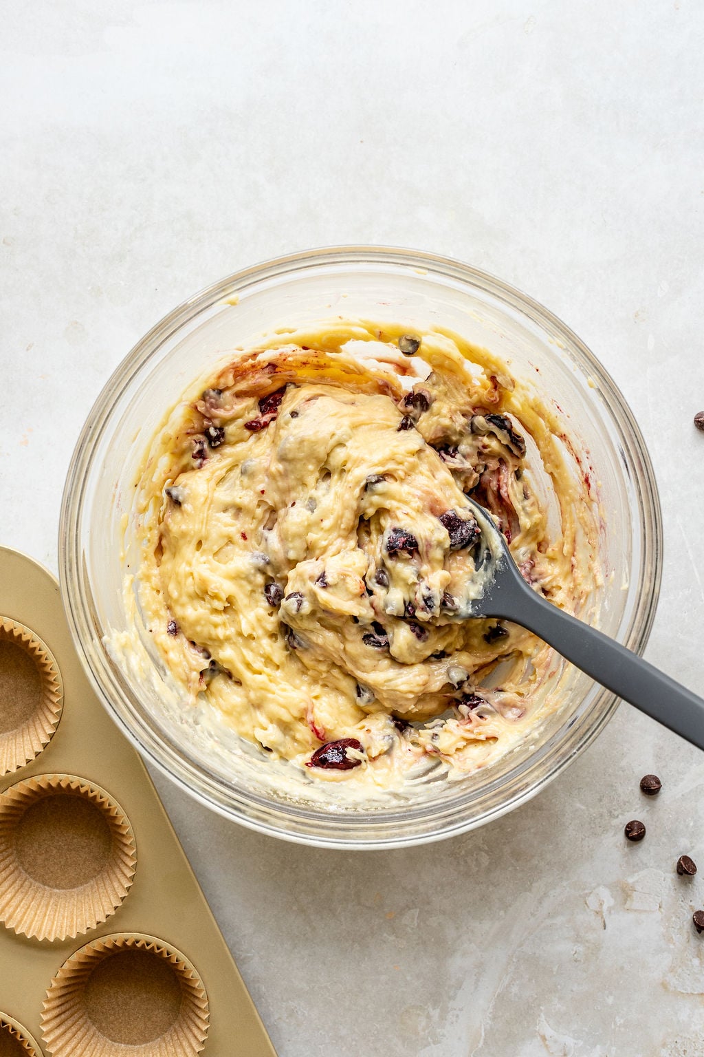 A glass bowl of muffin batter with chocolate chips and chopped cherries being mixed with a spatula, next to a muffin tin lined with paper cups.