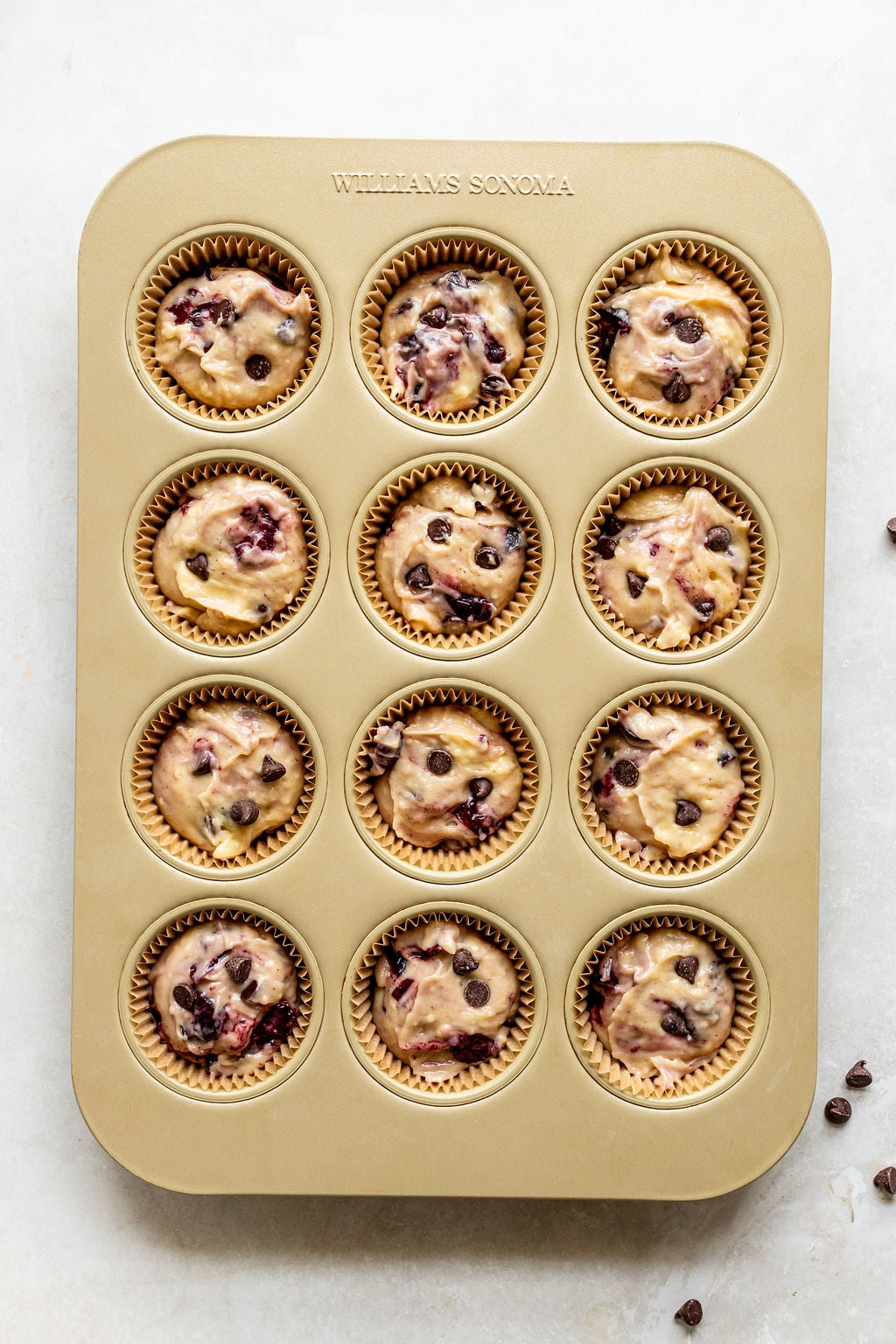 A gold muffin tin holds twelve unbaked muffins in paper liners, filled with batter containing chocolate chips and cherries.