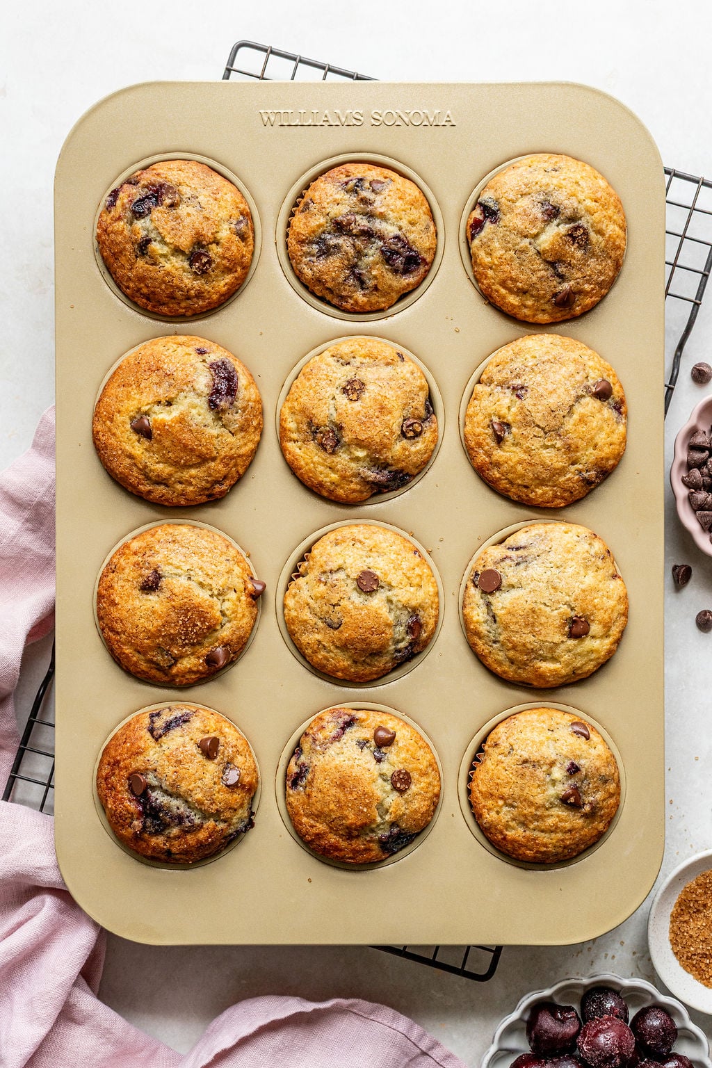 A muffin tin holds twelve baked muffins with visible chocolate chips and cherries, resting on a wire rack with a pink cloth nearby.