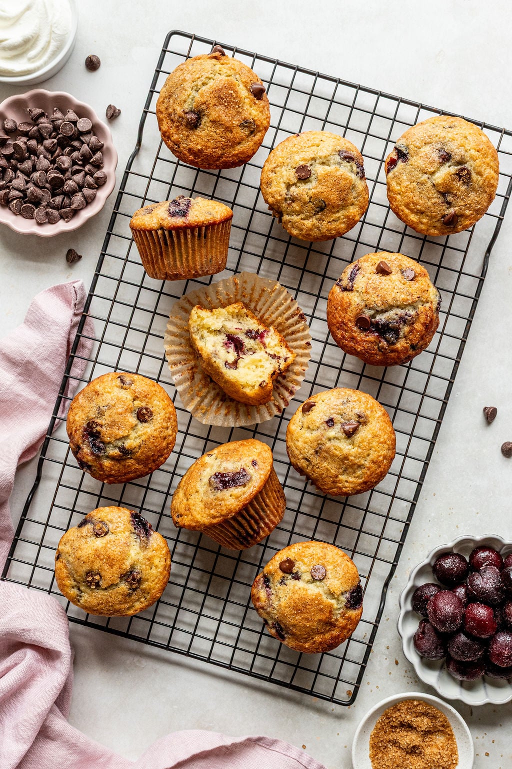 Twelve chocolate chip and cherry muffins on a cooling rack, with one muffin cut in half. Surrounding the rack are bowls of chocolate chips, whipped cream, cherries, and brown sugar.