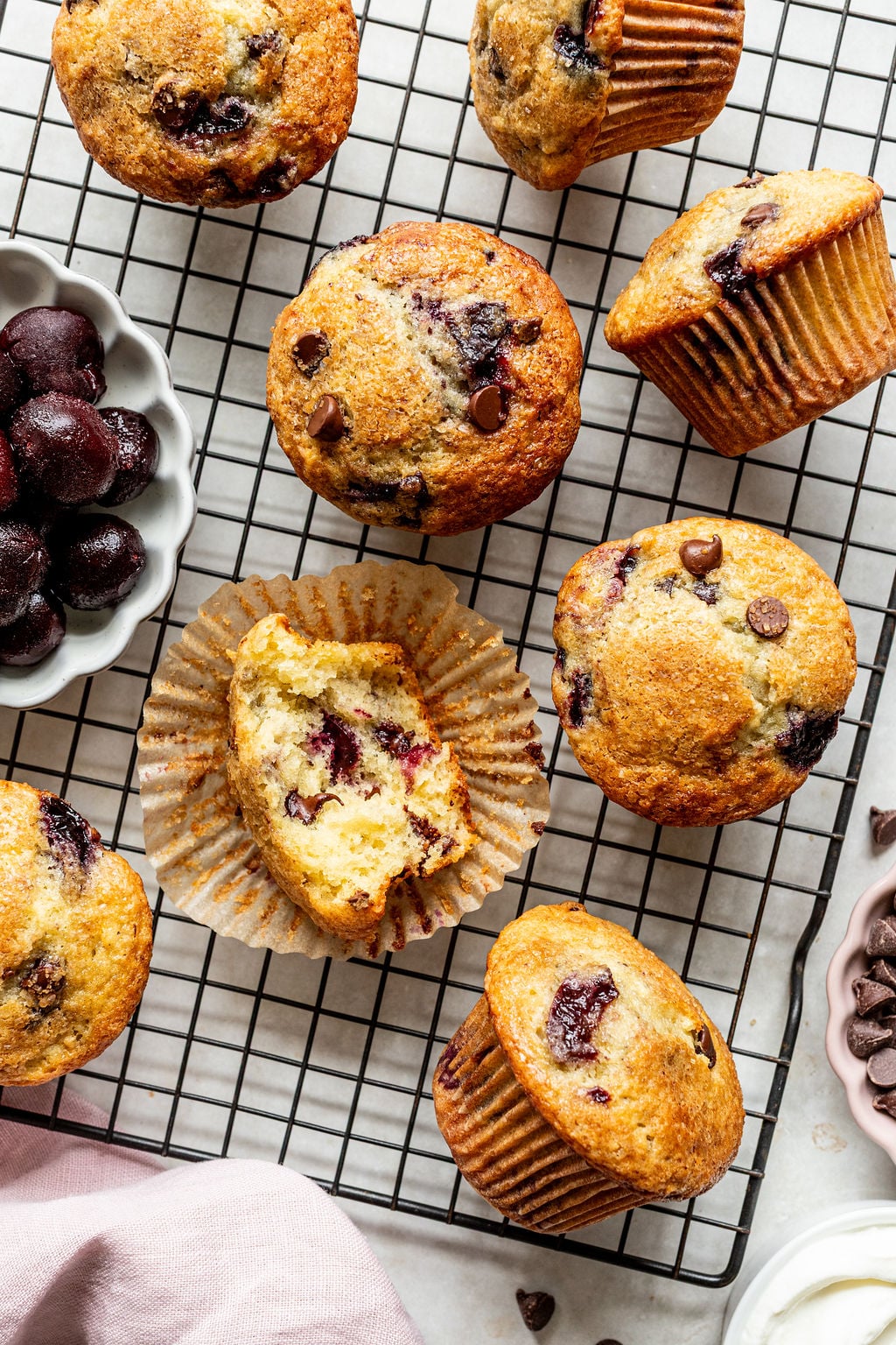 Chocolate chip cherry muffins on a cooling rack, with one muffin cut in half to show the inside, a small bowl of cherries, and chocolate chips nearby.