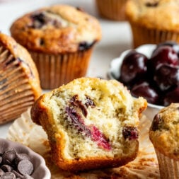 A close-up of a partially eaten muffin with visible cherries and chocolate chips, surrounded by whole muffins, a bowl of cherries, and a dish of chocolate chips on a white surface.