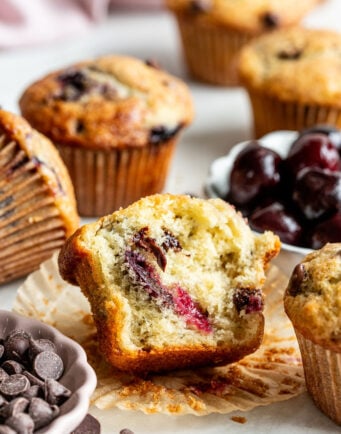 A close-up of a partially eaten muffin with visible cherries and chocolate chips, surrounded by whole muffins, a bowl of cherries, and a dish of chocolate chips on a white surface.