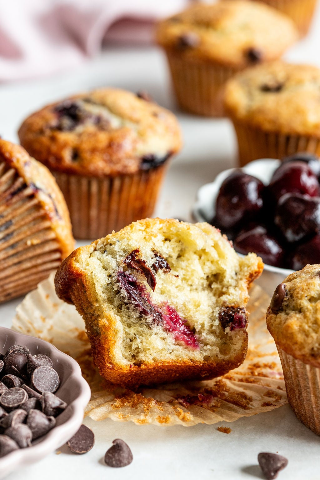 A close-up of a partially eaten muffin with visible cherries and chocolate chips, surrounded by whole muffins, a bowl of cherries, and a dish of chocolate chips on a white surface.