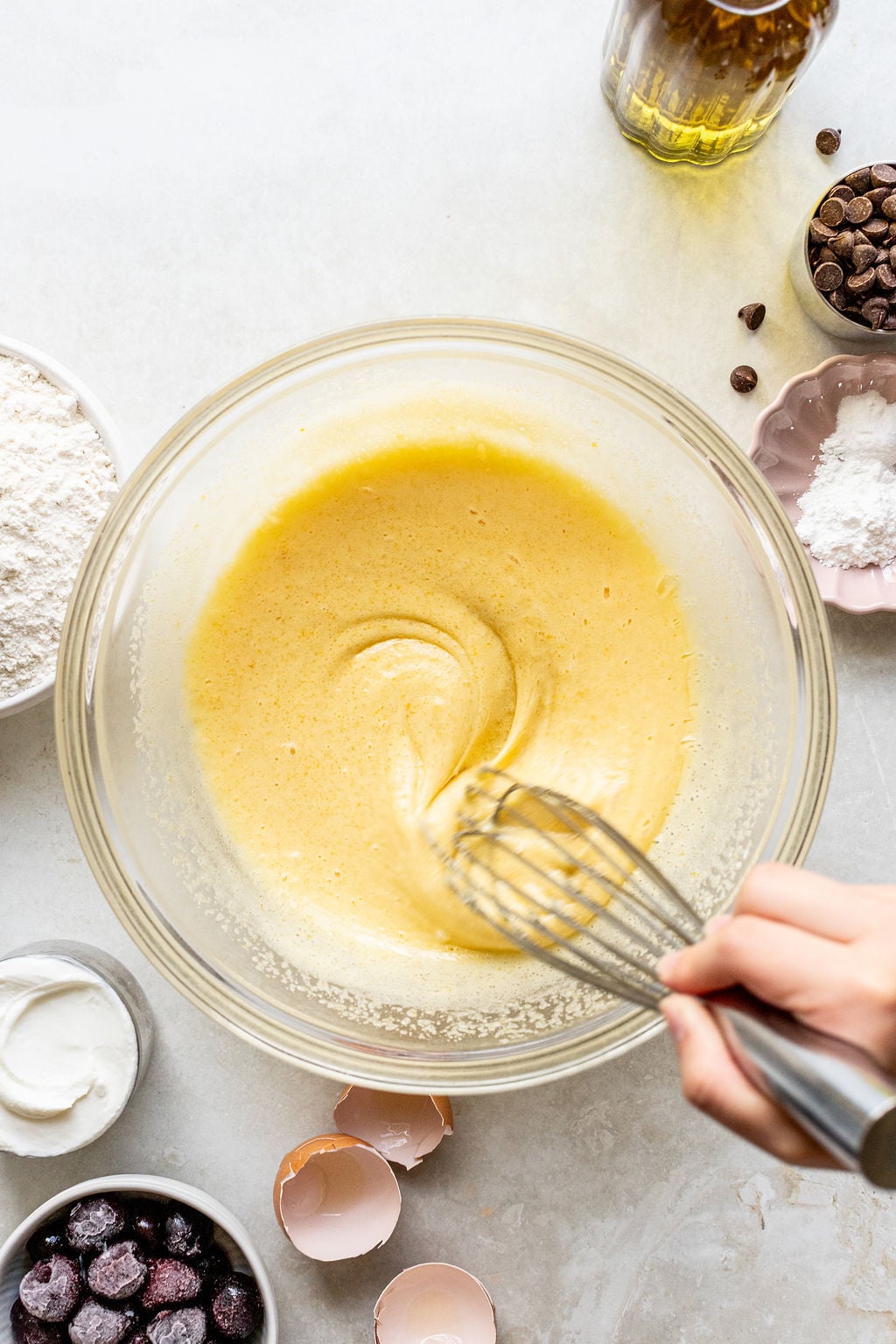 A hand whisking yellow batter in a glass bowl, surrounded by ingredients including flour, eggshells, cherries, chocolate chips, and a glass bottle.