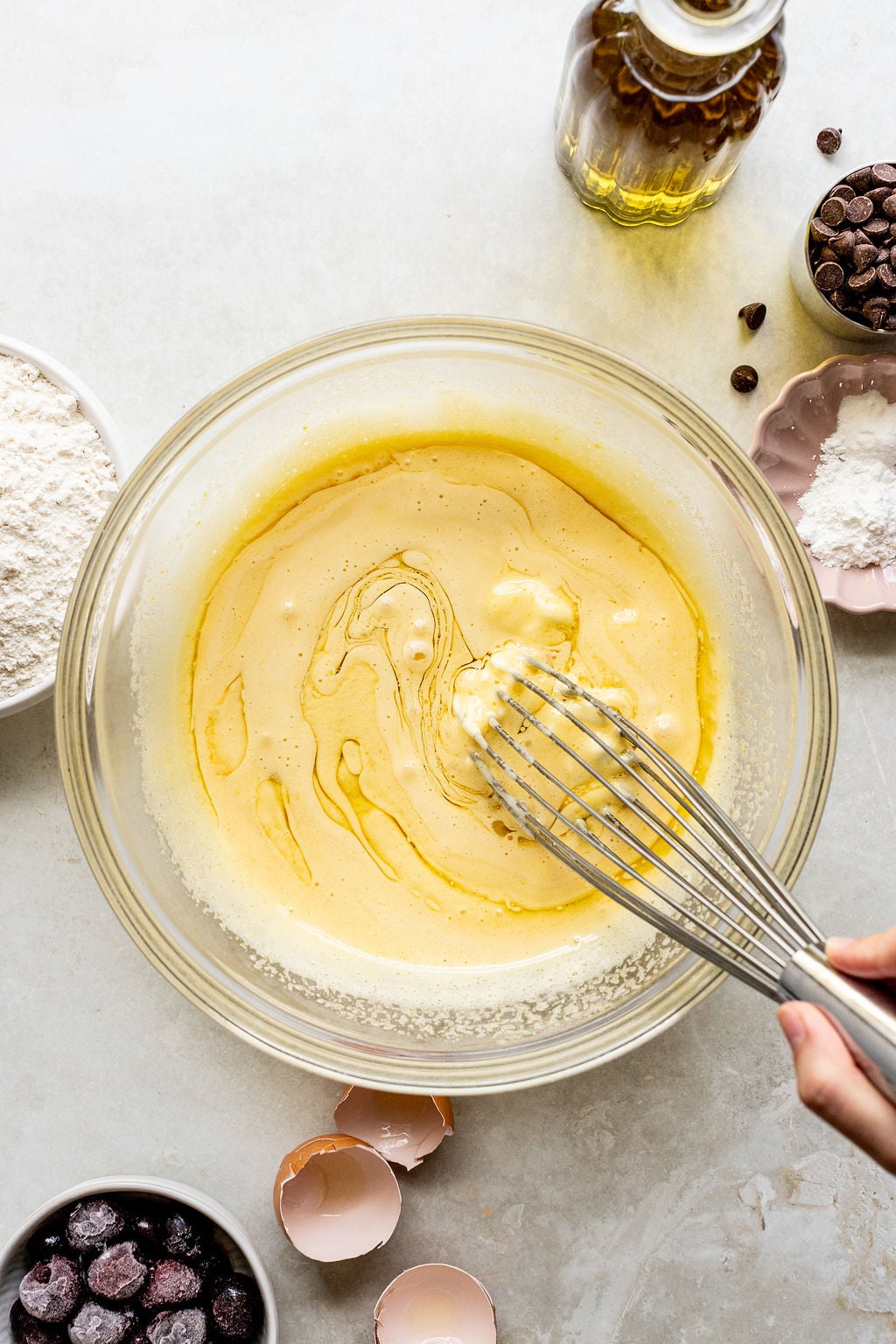 A hand whisking a yellow batter in a glass bowl, surrounded by flour, chocolate chips, cherries, egg shells, and a bottle of oil on a light countertop.