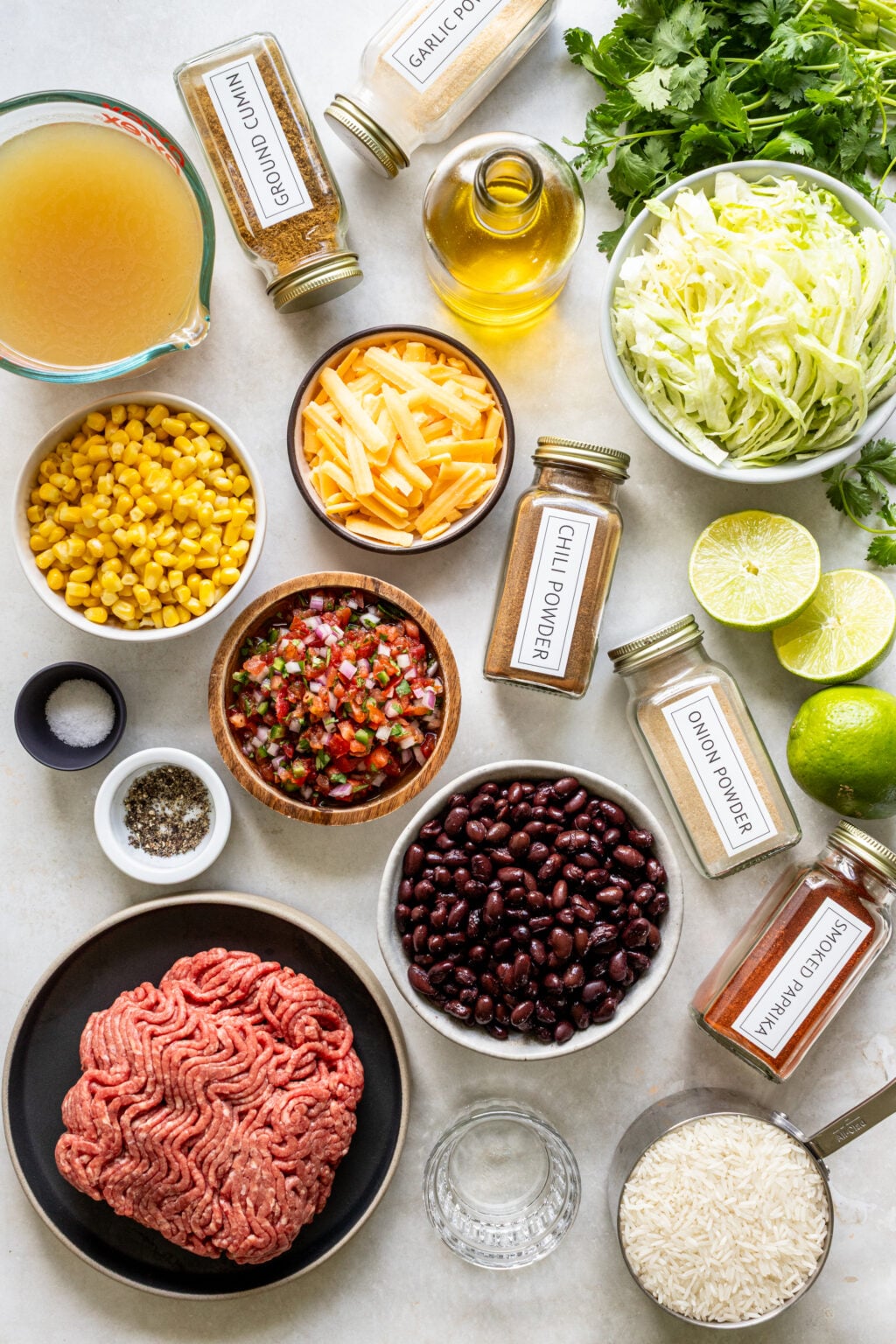 Ingredients for a taco-inspired dish arranged on a countertop, including ground beef, black beans, corn, cheese, spices, broth, oil, rice, shredded lettuce, pico de gallo, cilantro, and limes.