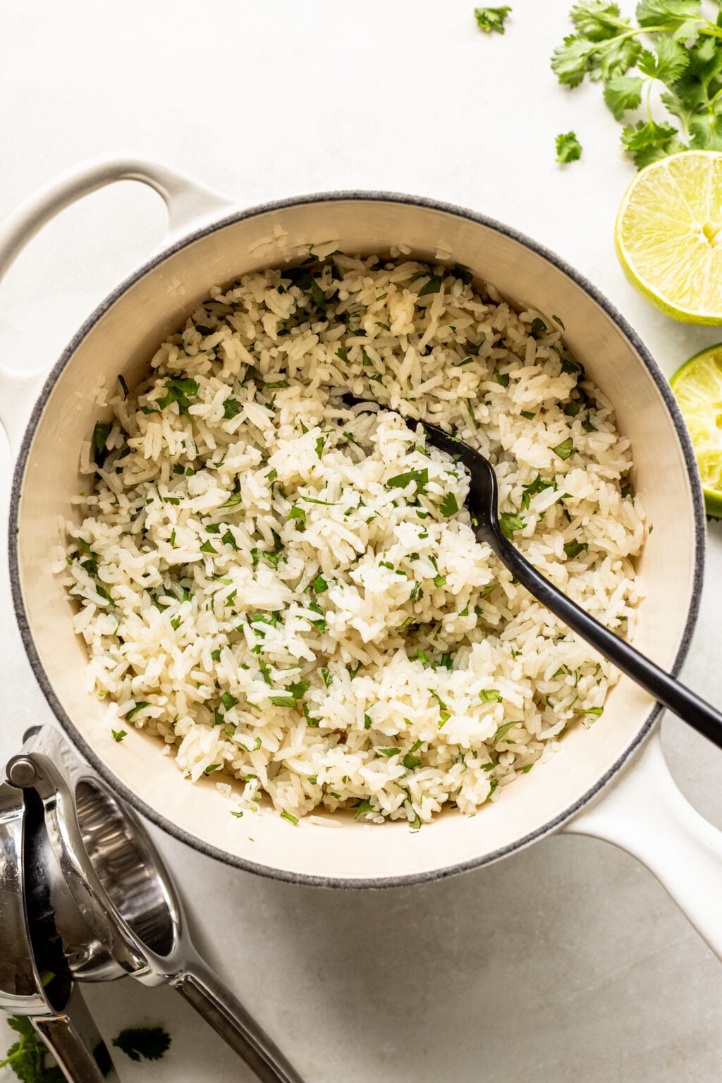 A white pot filled with cooked rice mixed with chopped cilantro, with a black serving spoon inside. Lime halves and cilantro leaves are on the side.