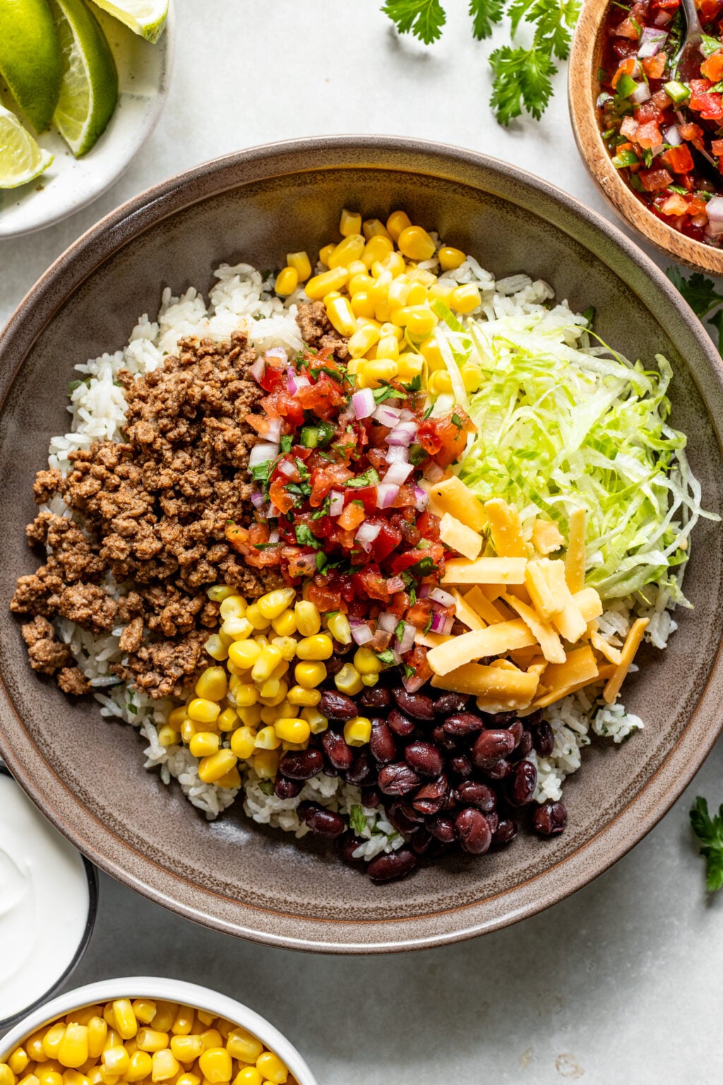 A bowl with rice, ground beef, corn, black beans, shredded lettuce, shredded cheese, and salsa, surrounded by small bowls of limes, pico de gallo, sour cream, and corn.