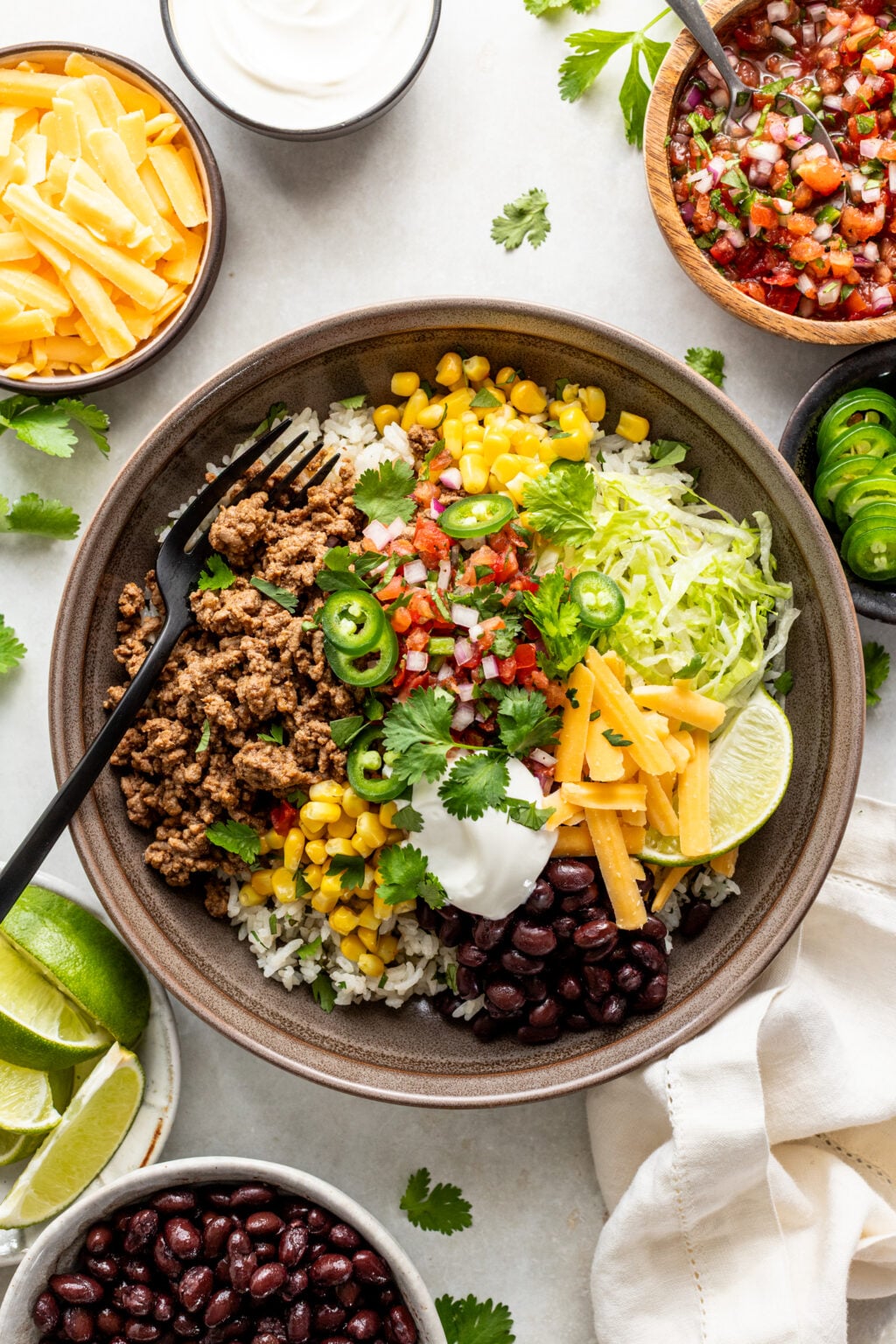 A burrito bowl with ground beef, rice, black beans, corn, shredded lettuce, cheese, sour cream, salsa, jalapenos, lime wedges, and cilantro, surrounded by small bowls of ingredients.