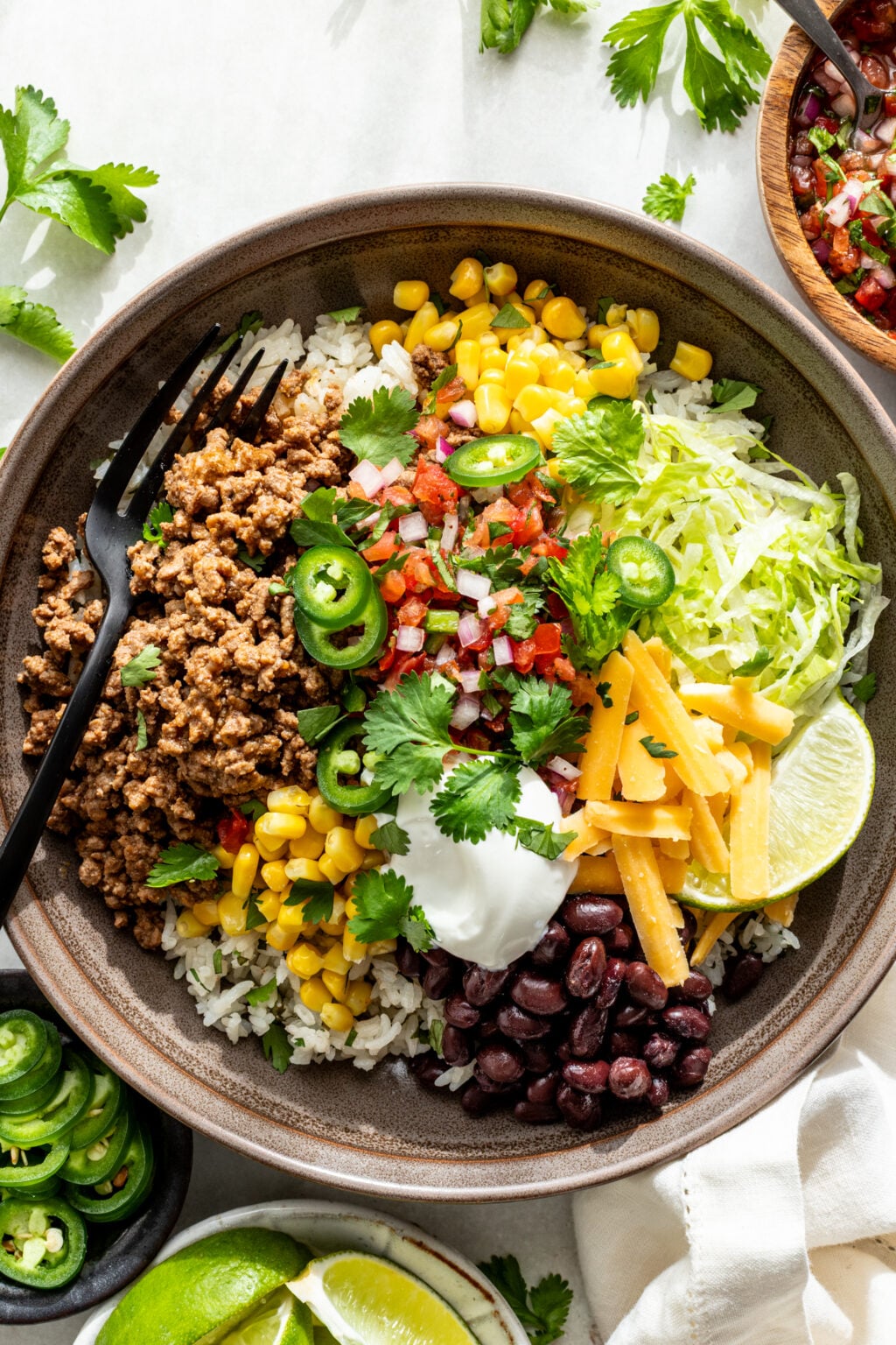 A bowl filled with rice, seasoned ground meat, black beans, corn, shredded lettuce, cheese, salsa, sour cream, jalape&ntilde;o slices, cilantro, and lime wedges, with a black fork.