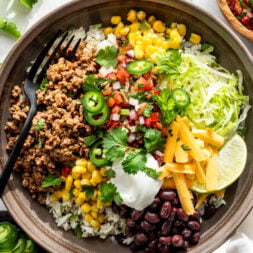 A bowl filled with rice, seasoned ground meat, black beans, corn, shredded lettuce, cheese, salsa, sour cream, jalape&ntilde;o slices, cilantro, and lime wedges, with a black fork.