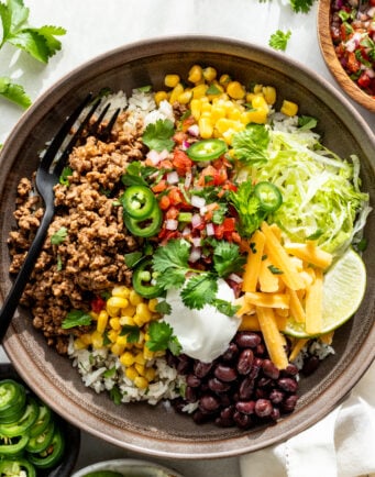 A bowl filled with rice, seasoned ground meat, black beans, corn, shredded lettuce, cheese, salsa, sour cream, jalapeño slices, cilantro, and lime wedges, with a black fork.