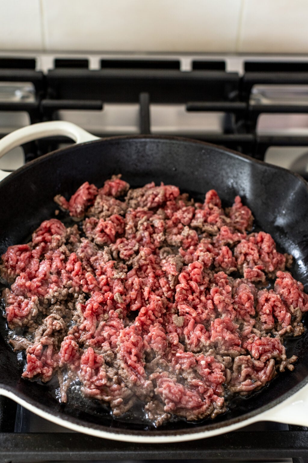 Ground beef cooking in a black cast iron skillet on a stovetop, with some parts browned and others still raw.