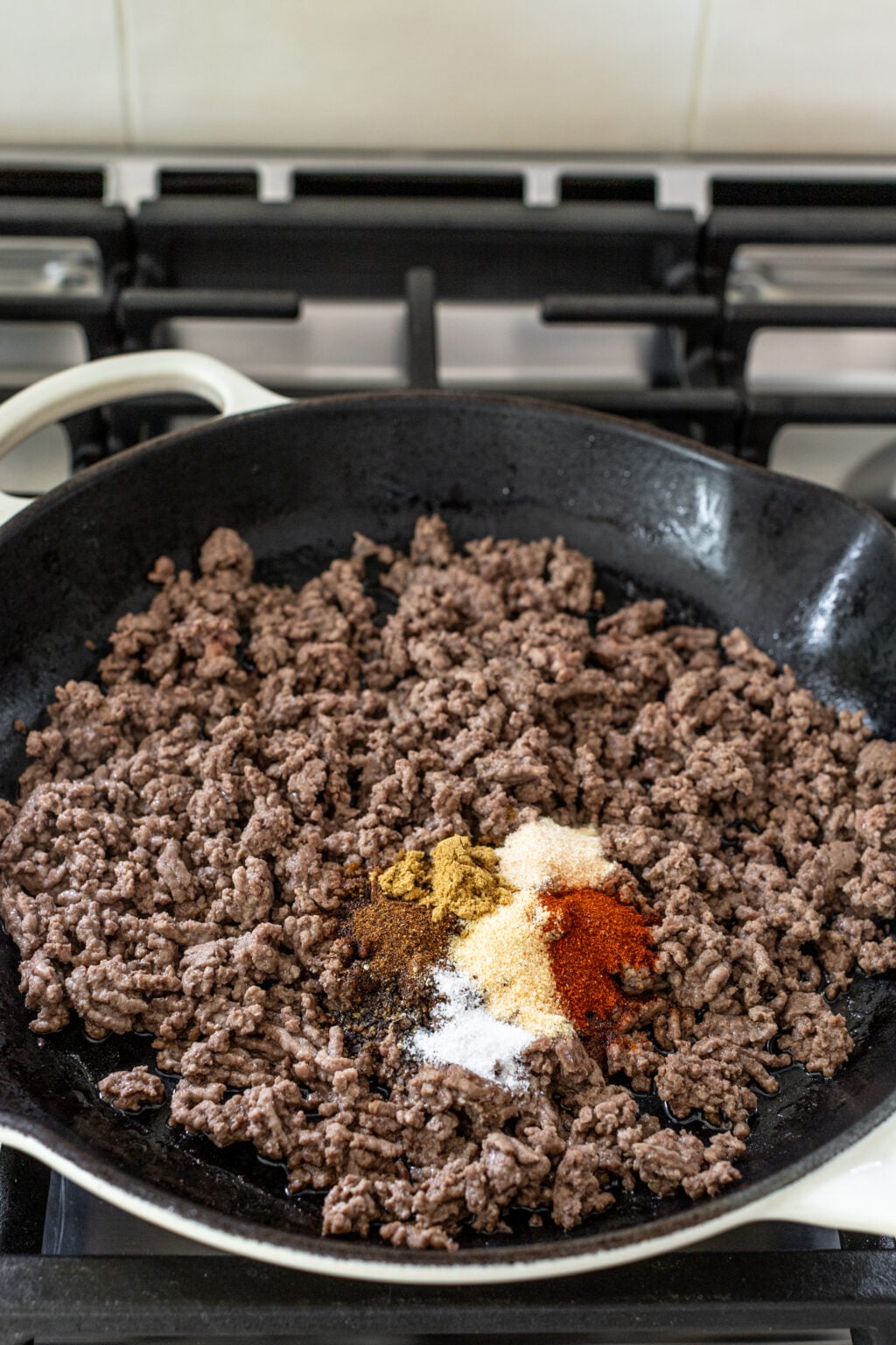 Ground beef cooking in a black skillet on a stove, with several different spices piled on top of the meat.