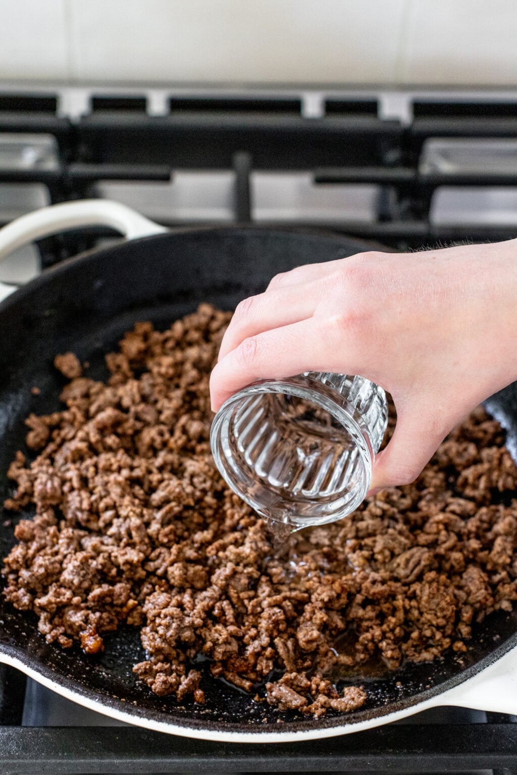 A hand pours water from a glass into a skillet filled with cooked ground beef on a stovetop.