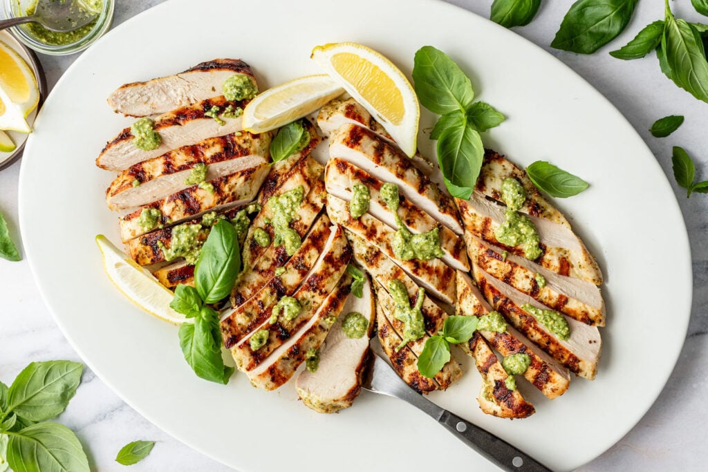 Grilled sliced chicken breast on a white platter, topped with green pesto sauce, garnished with fresh basil leaves and lemon wedges. A fork is placed on the plate.