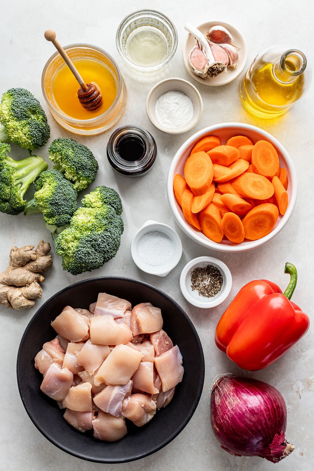 Raw chicken pieces, sliced carrots, broccoli, red bell pepper, red onion, ginger, garlic, honey, soy sauce, oil, vinegar, salt, pepper, and cornstarch arranged on a countertop.
