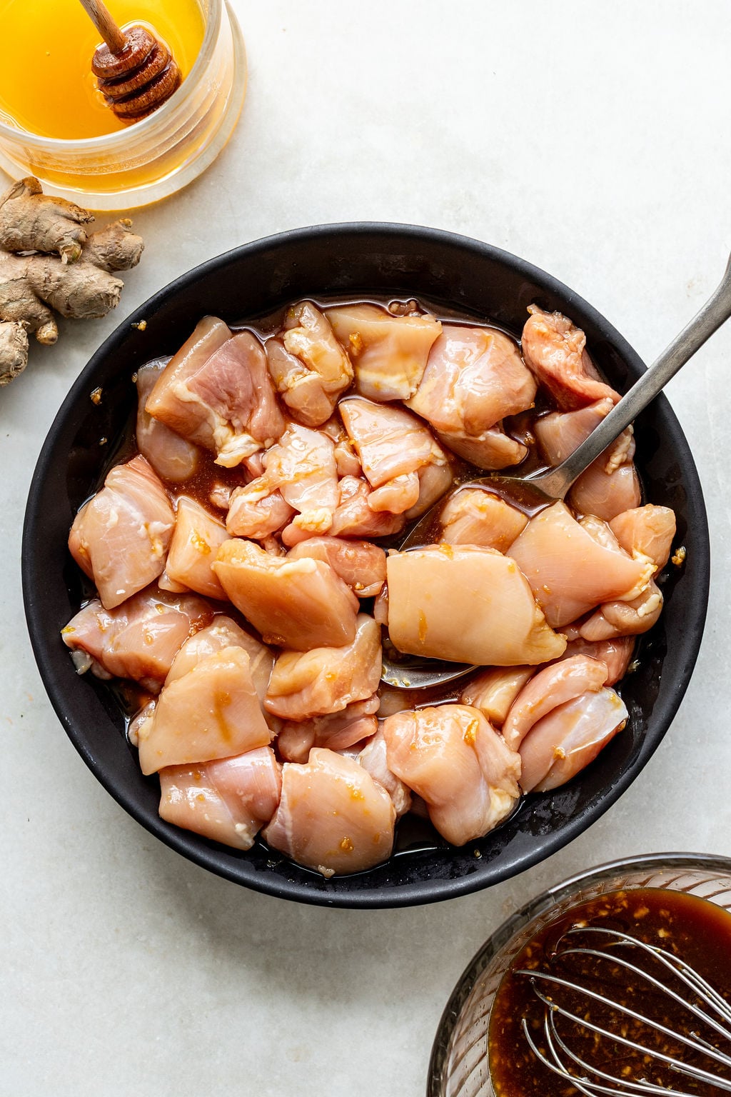 A bowl of raw, marinated chicken pieces with a spoon, surrounded by fresh ginger, a jar of honey with a dipper, and a bowl of marinade on a white surface.