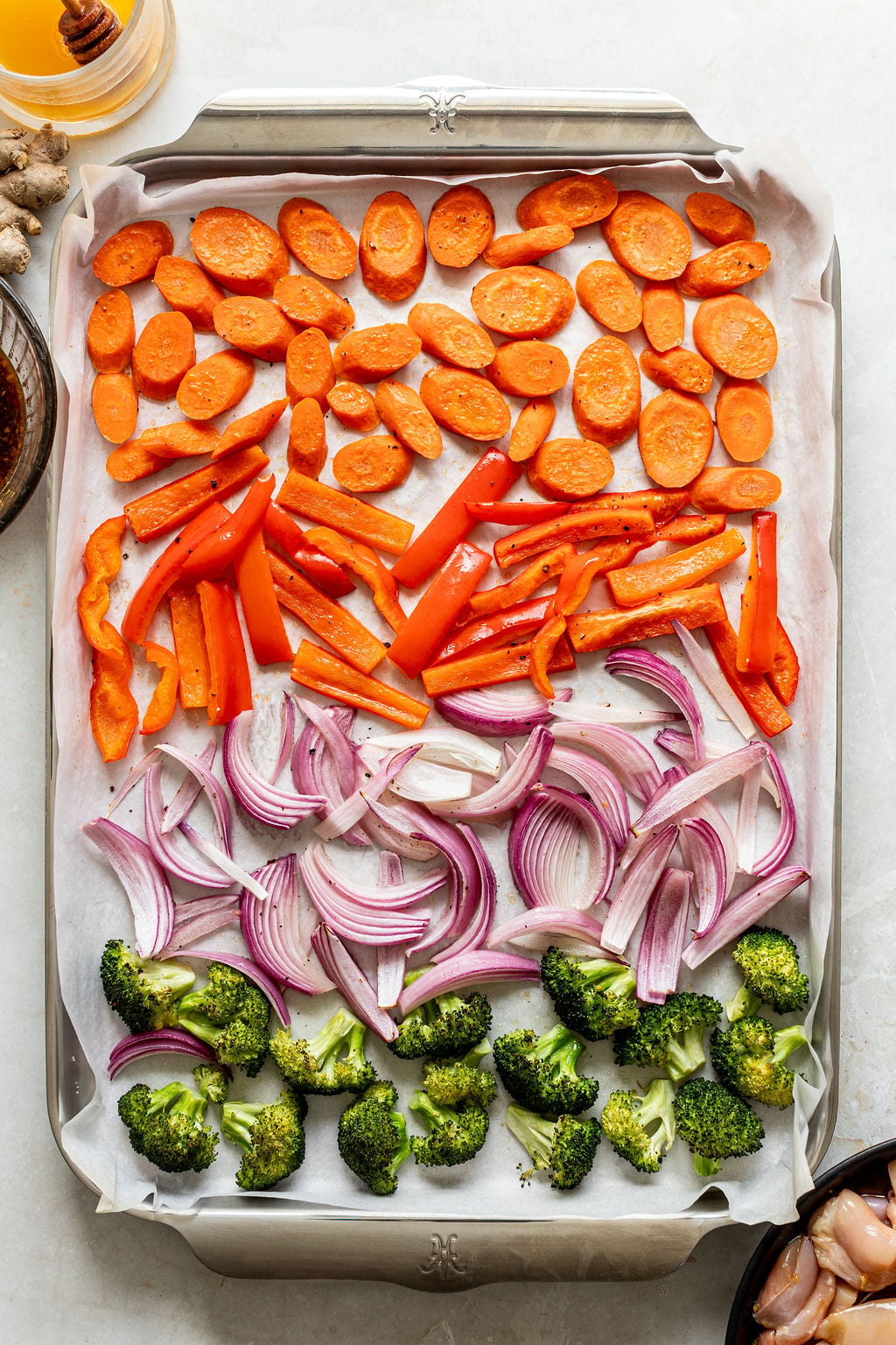 A baking sheet lined with parchment paper displays rows of sliced carrots, red bell peppers, red onions, and broccoli florets, ready for roasting.