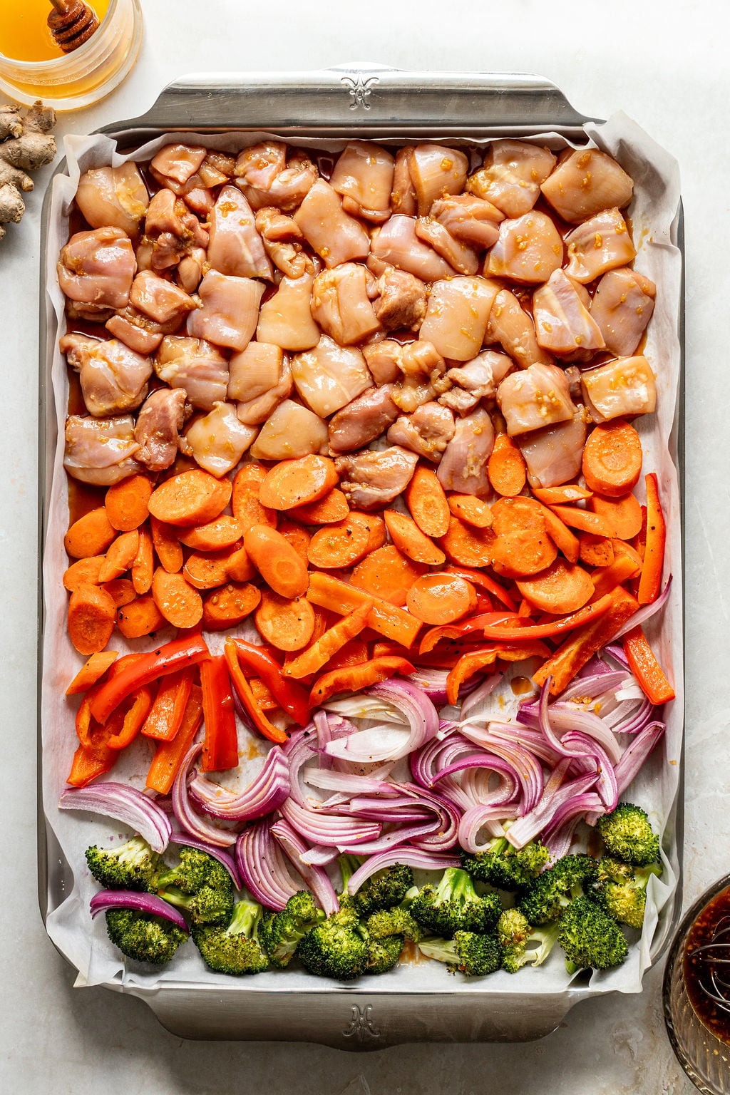 Sheet pan with rows of raw diced chicken, sliced carrots, red bell peppers, red onions, and broccoli on parchment paper.