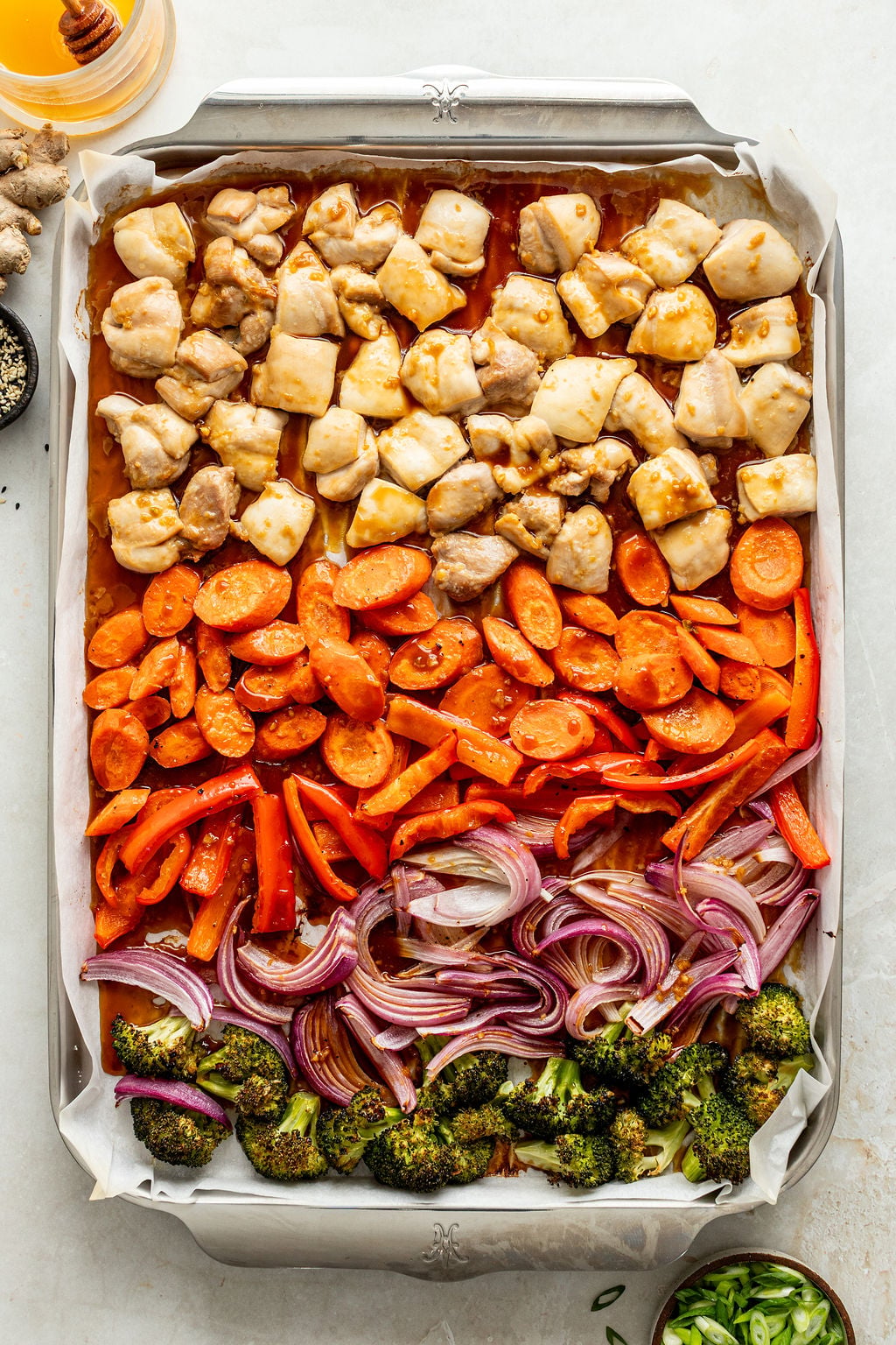 Sheet pan with rows of diced chicken, sliced carrots, red bell pepper, red onion, and broccoli, all seasoned and ready for roasting.