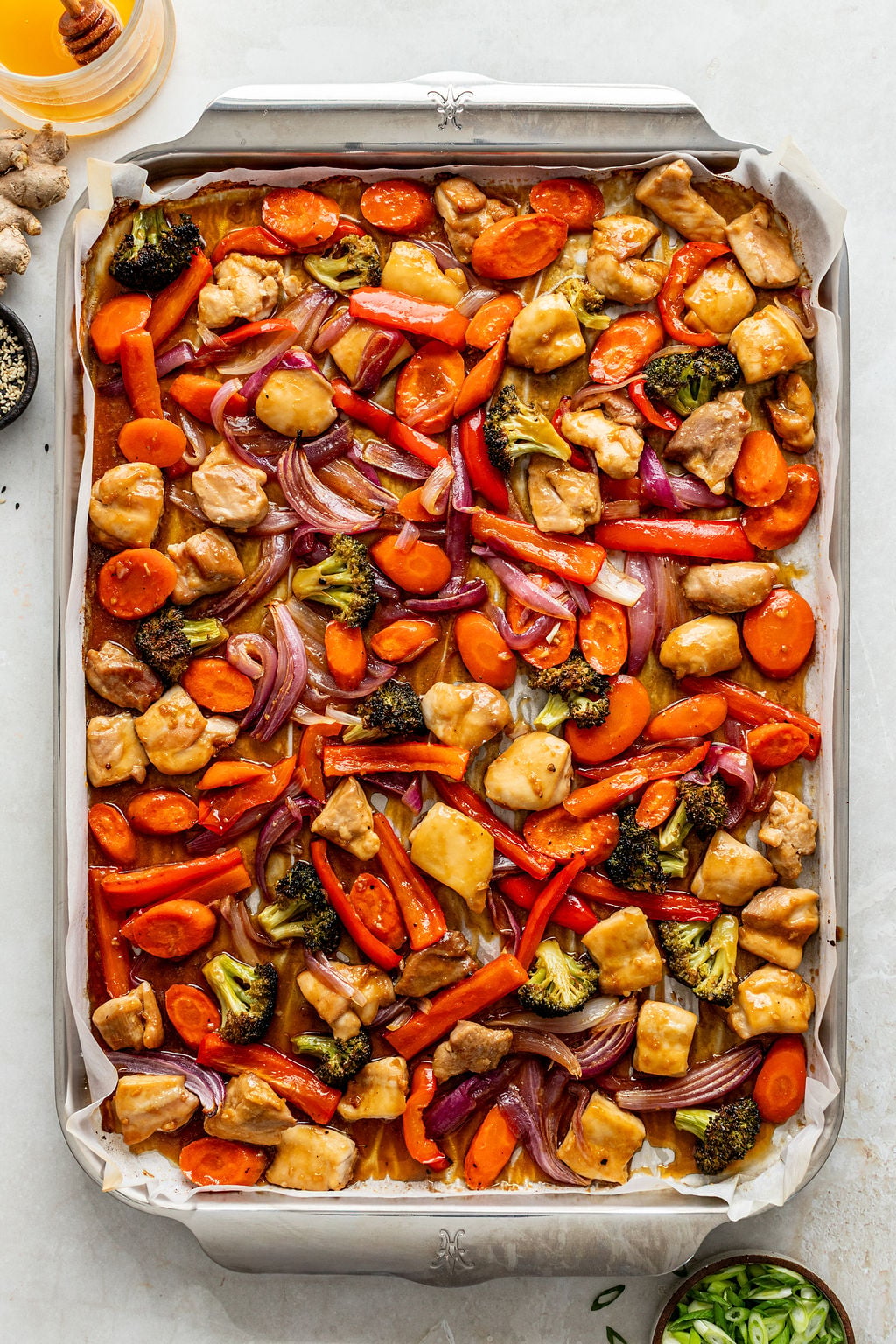 A sheet pan lined with parchment paper holds baked chicken pieces, sliced carrots, red bell peppers, red onions, and broccoli in a light sauce.